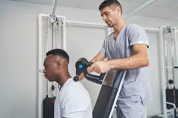 Physical therapist applying pressure on a man's upper back with a massage gun 